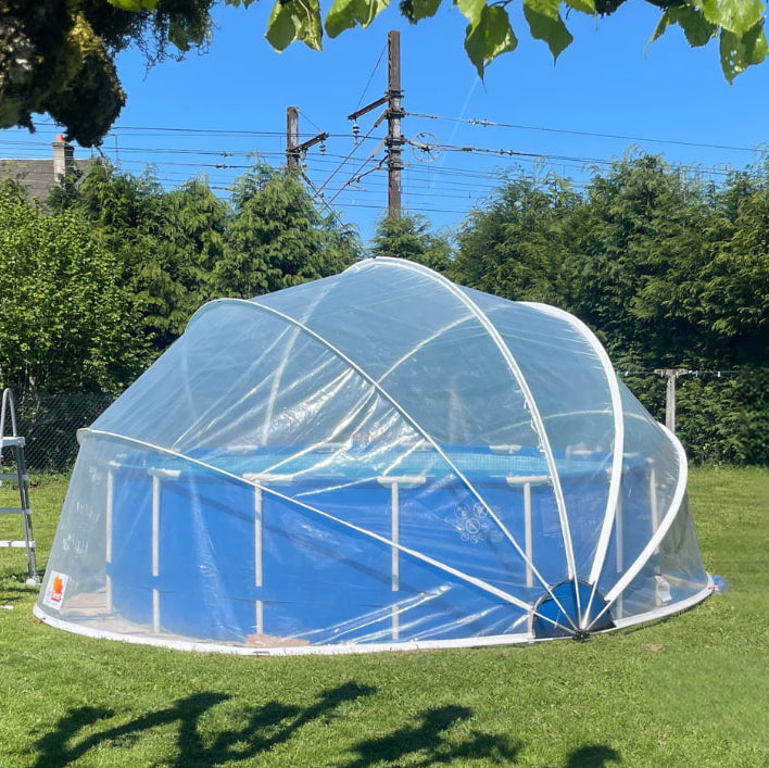 Clear pool cover over an above-ground pool in a grassy area with trees and power lines in the background.
