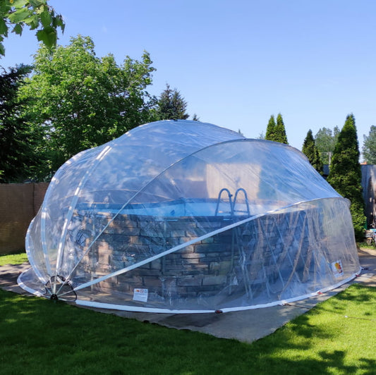 A clear, oval-shaped pool dome covering a pool area, set up on a grassy backyard with trees in the background.