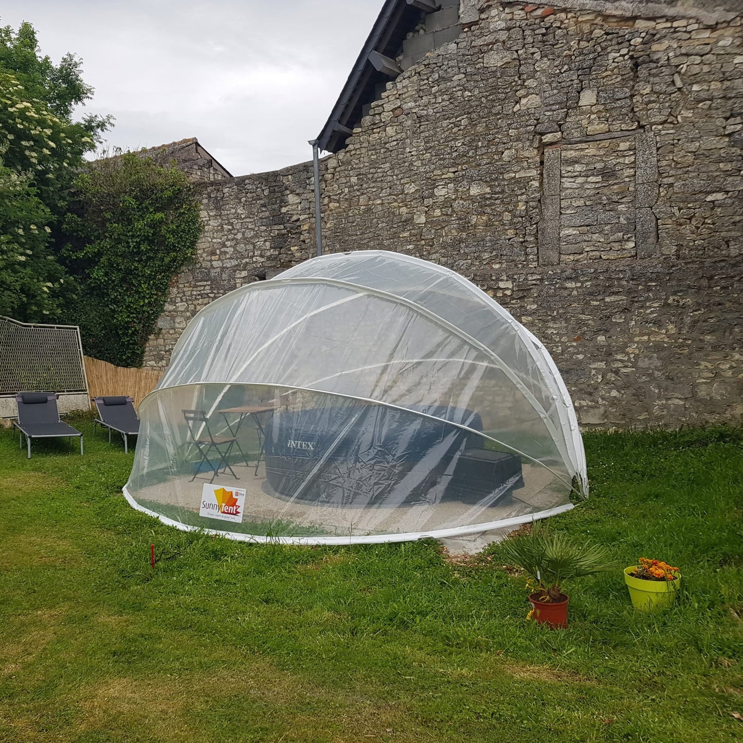 A clear, round pool dome tent set up on a grassy area with a stone wall in the background and chairs to the side.